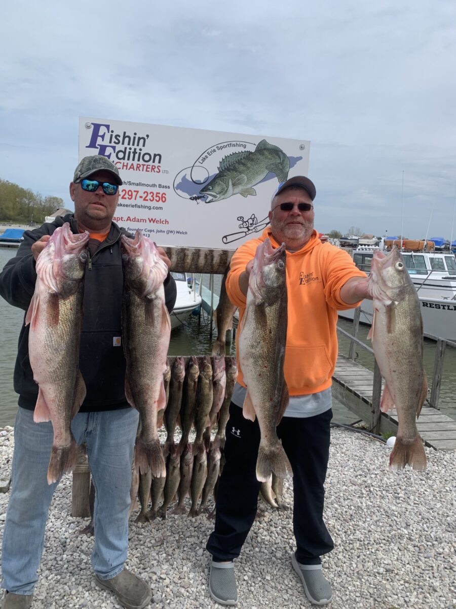 Two men displaying their catch of large fish in front of a charter sign.
